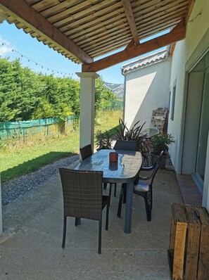 Un patio exterior con mesa y sillas, una pérgola arriba y vistas a un paisaje verde.