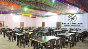 A large dining hall with multiple tables and chairs, a patterned ceiling, and a logo for Ozaki Jaisalmer. - Ozaki Desert Camp Jaisalmer  (Jaisalmer)