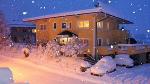 A two-story house with a snow-covered roof and driveway, surrounded by snow-covered bushes and trees. - Winter dream vacation apartment in the Zillertal in direct proximity to the Horbergbahn ski lift (Schwendau)
