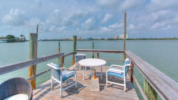 A wooden deck with a table and chairs, overlooking a calm body of water with distant buildings. - Front-Row Waterfront Escape – 1BR Condo on Clearwater Beach (Clearwater Beach)