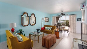 A living room with a yellow sofa, a brown armchair, a glass coffee table, and a patterned ottoman. There is a dining area with a table and chairs, a ceiling fan, and a view through the window. - Front-Row Waterfront Escape – 1BR Condo on Clearwater Beach (Clearwater Beach)