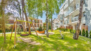 A playground with swings and a slide, surrounded by trees and a building with balconies. - Apartamenty Sun & Snow Nautikka Park (Krynica Morska)