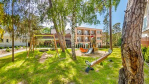 A playground with swings and a slide, surrounded by trees and a building with balconies in the background. - Apartamenty Sun & Snow Nautikka Park (Krynica Morska)