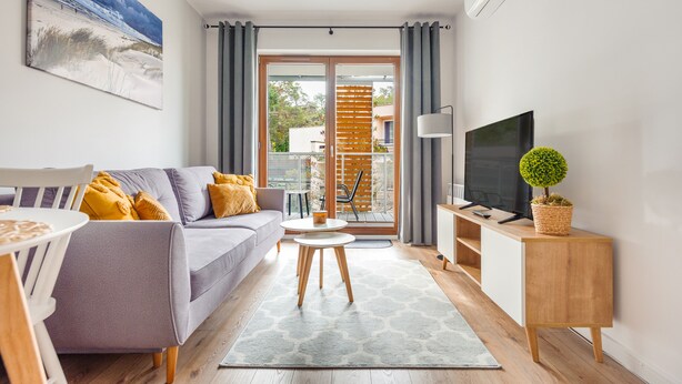 A modern living room with a grey sofa, a small round table, a TV on a wooden stand, and a balcony with a view of the outdoors. - Apartamenty Sun & Snow Nautikka Park (Krynica Morska)