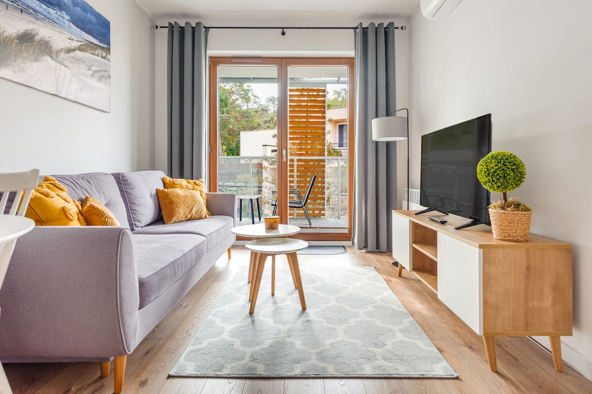 A modern living room with a grey sofa, a small round table, a TV on a wooden stand, and a balcony with a view of the outdoors.