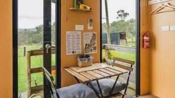 A room with a wooden table and two chairs, a window with a view of greenery, and a wall-mounted shelf with items.