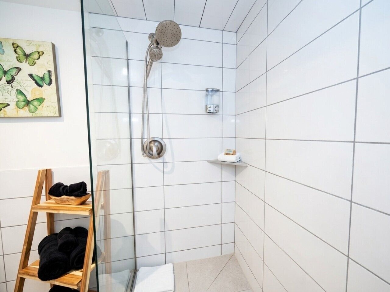 A bathroom with white tiled walls, a glass shower enclosure, a wooden shelf with towels, and a small shelf with toiletries.
