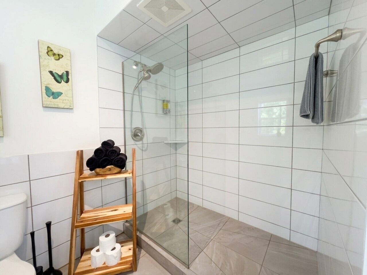 A modern bathroom with a glass shower enclosure, white tiled walls, and a wooden shelf with towels and toiletries.