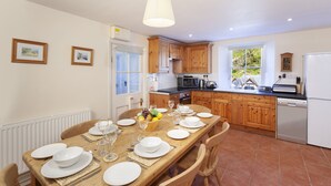 A dining area with a wooden table set for four, kitchen cabinets, a refrigerator, and a window with a view of greenery. - Lorna Doone Cottage, Lynmouth (Lynmouth)