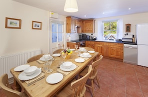 A dining area with a wooden table set for four, kitchen cabinets, a refrigerator, and a window with a view of greenery.