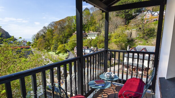 A balcony with a view of a hillside, a table with chairs, and a red cushion. - Lorna Doone Cottage, Lynmouth (Lynmouth)