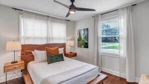 A bedroom with a bed, a ceiling fan, two bedside tables with lamps, a bench, and a window with curtains. - Modern 2 BR Condo - 2 block from San Marco (Jacksonville)