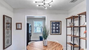 A dining area with a round wooden table, chairs, and a potted plant. There are framed artworks on the walls and a metal shelving unit with items on it.