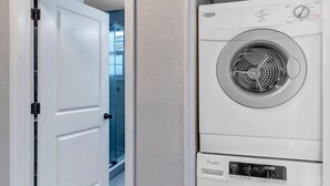 A laundry room with a stacked washer and dryer, a white door leading to a hallway, and a wooden floor.