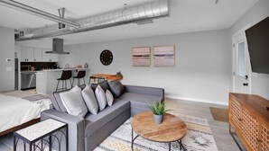 A modern living room with a grey sofa, a round wooden coffee table, and a TV on the right. There is a kitchen area with a dining table and chairs in the background. - Chic & modern studio in the heart of San Marco! (Jacksonville)