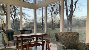 A conservatory with a wooden table and chairs, a leopard-print armchair, and a view of trees and a fence.