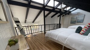 A bedroom with a wooden floor, a bed with white bedding, a small plant on a table, and a staircase leading to an outdoor area.