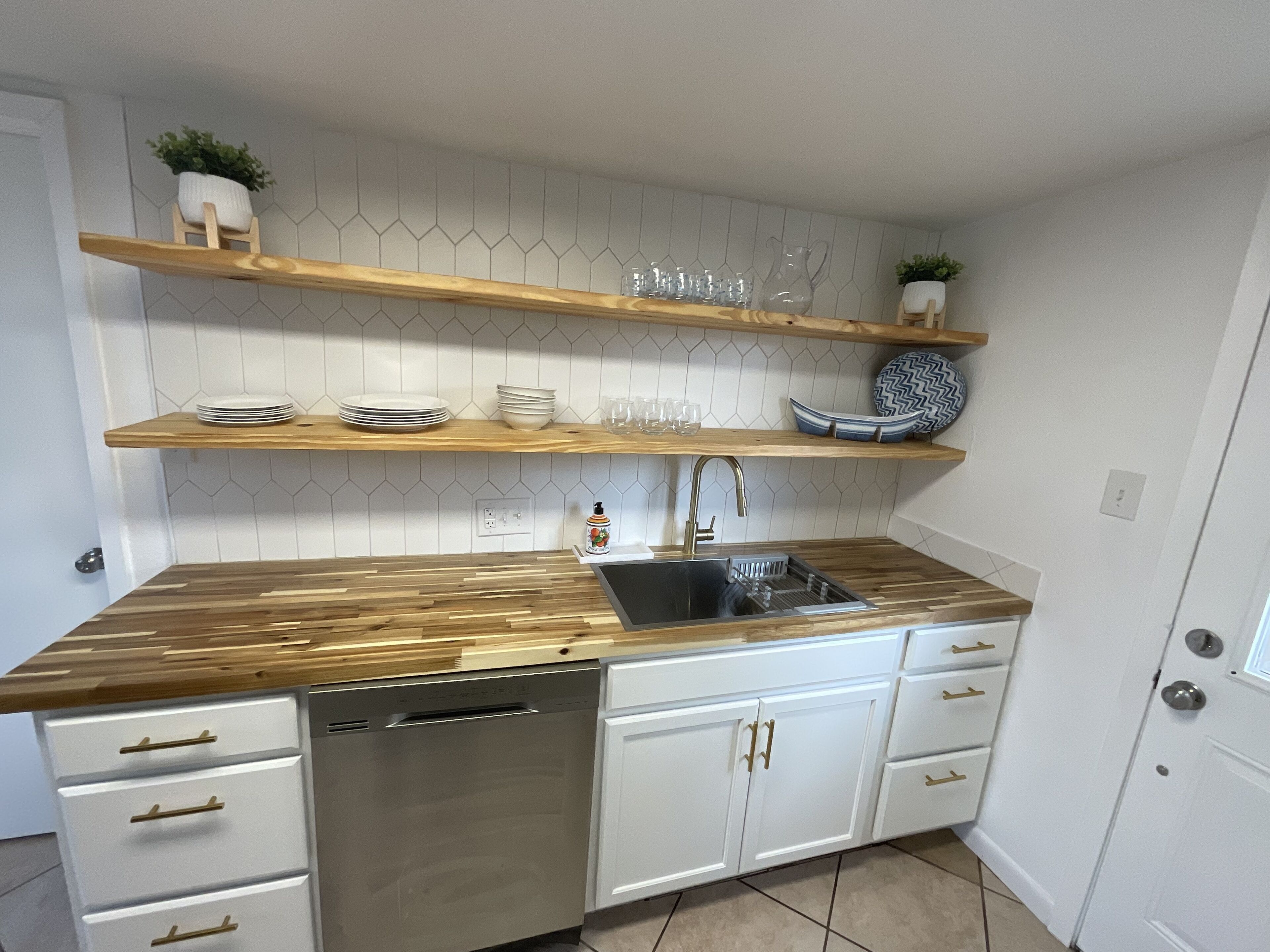 A kitchen with wooden countertops, white cabinets, and a stainless steel dishwasher. There are two wooden shelves above the sink, holding dishes and glasses.