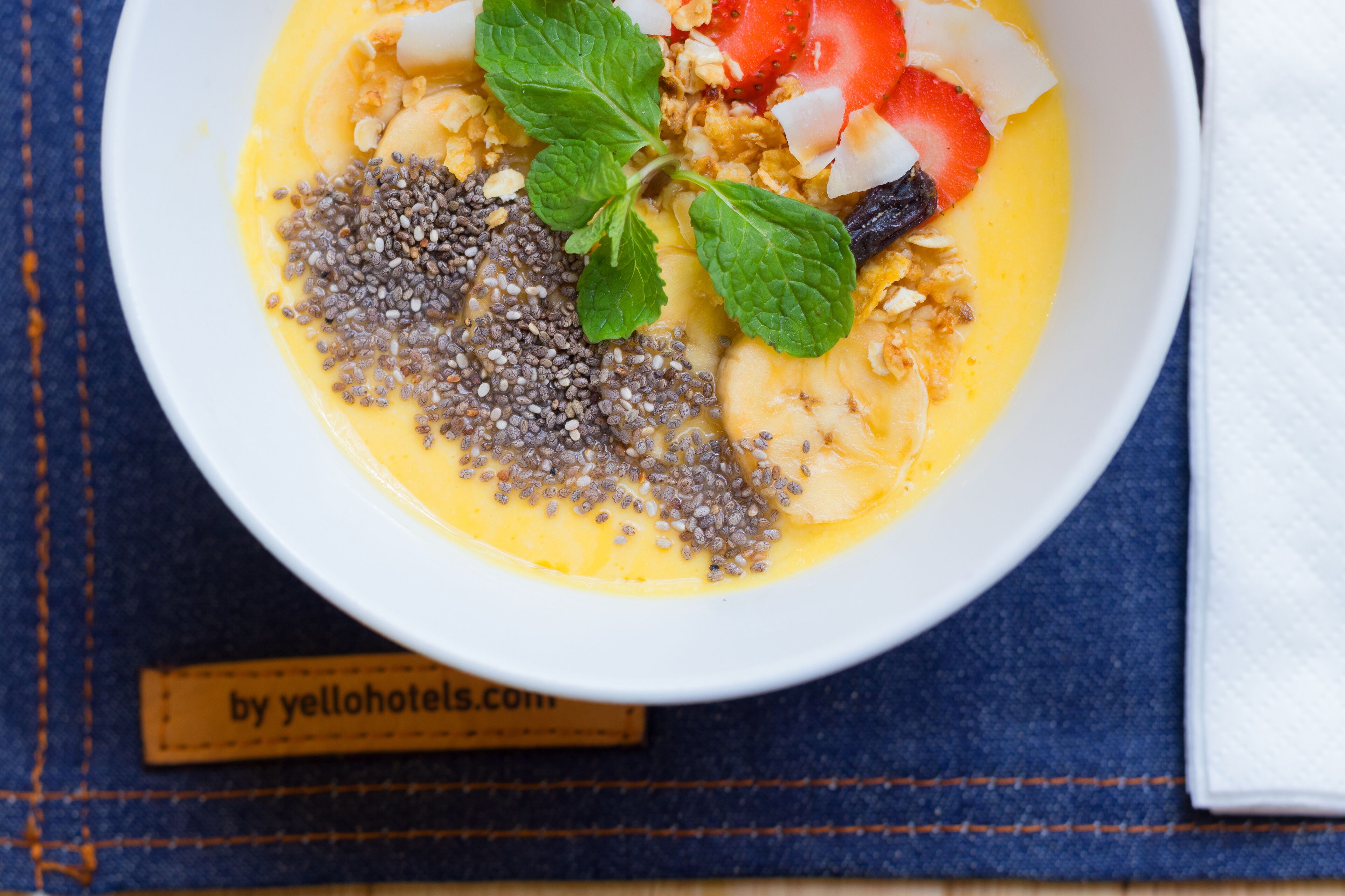 Un tazón de comida con semillas de chía, fresas, rodajas de plátano y una hoja de menta sobre un paño azul.