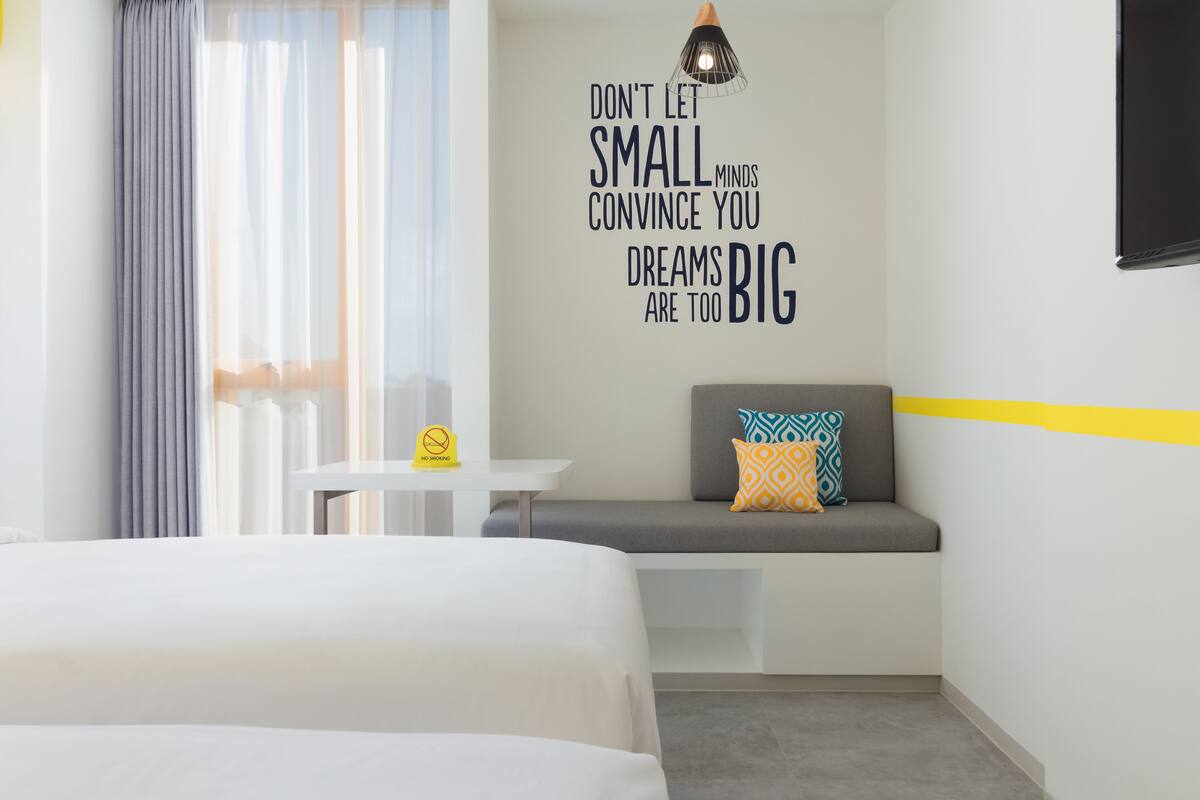 A modern hotel room with a bed, a small table, a bench with cushions, and a motivational quote on the wall.