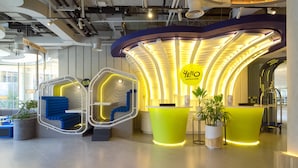 Uma área de recepção de hotel moderna, com um balcão amarelo curvo, um espaço de estar azul e uma placa indicando NETZONE.