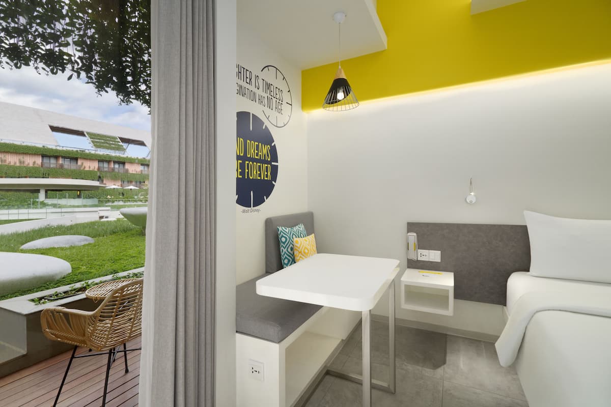 A modern hotel room with a view of a building with green roofs, a small table, a bench, and a bed.
