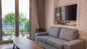 Un salon avec un canapé, un téléviseur fixé au mur en briques, un balcon avec vue sur la mer et une petite table.