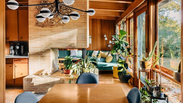 Un salon chaleureux avec une cheminée en briques, des poutres apparentes en bois au plafond et de grandes fenêtres donnant sur l’extérieur.