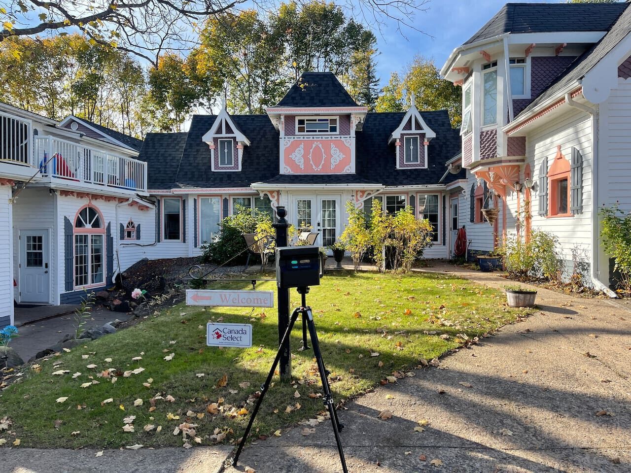 A large, multi-story house with a prominent 'Welcome' sign, a Canada Select sign, and a security camera on a tripod in the driveway.