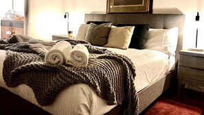 Een netjes opgemaakt bed met opgerolde handdoeken, een bruin gebreid plaid en een schilderij erboven.
