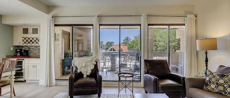 Sala de estar com sofá, cadeiras, mesa de centro e vista para um pátio externo através de amplas portas de vidro.