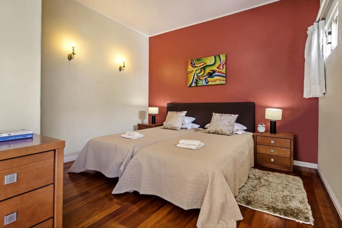 A hotel room with two beds, a wooden dresser, a red accent wall, and a painting.