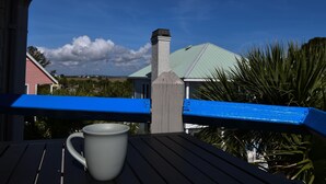 A white mug on a wooden table with a blue railing, overlooking a house with a green roof and a chimney. - Sandy Toes Condo's (Saint Helena Island)