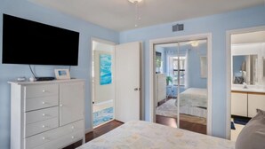 A bedroom with a bed, a TV mounted on the wall, a dresser, and a door leading to another room. - Sandy Toes Condo's (Saint Helena Island)