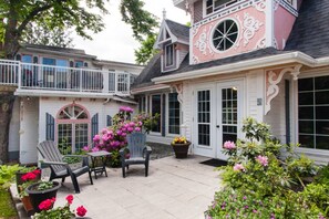 A two-story house with a balcony, a patio with chairs and potted plants, and a large window.