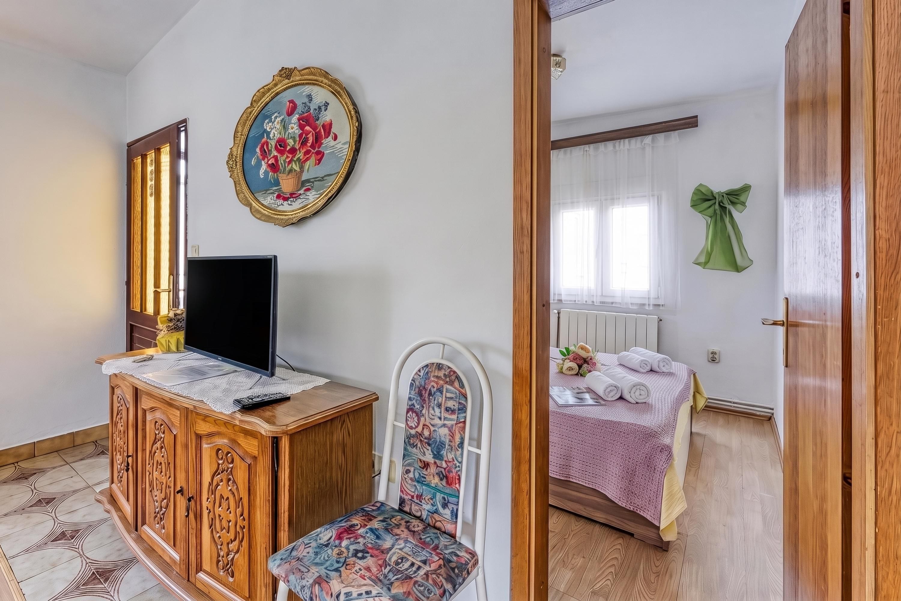 A room with a wooden cabinet, a television, a chair with a floral seat, a bed with a pink bedspread, and a green bow decoration on the wall.