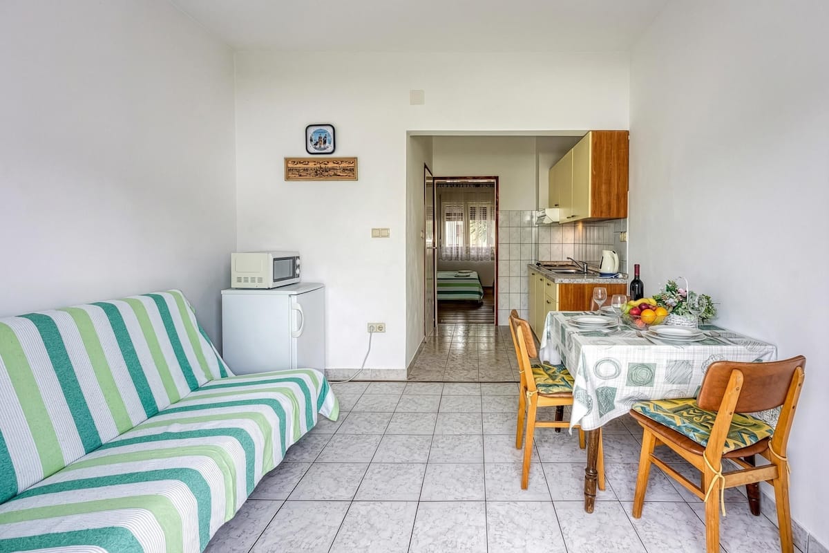 Un espace de vie compact comprenant un canapé à rayures, une table à manger avec des chaises, un petit coin cuisine et une porte visible donnant sur une autre pièce.