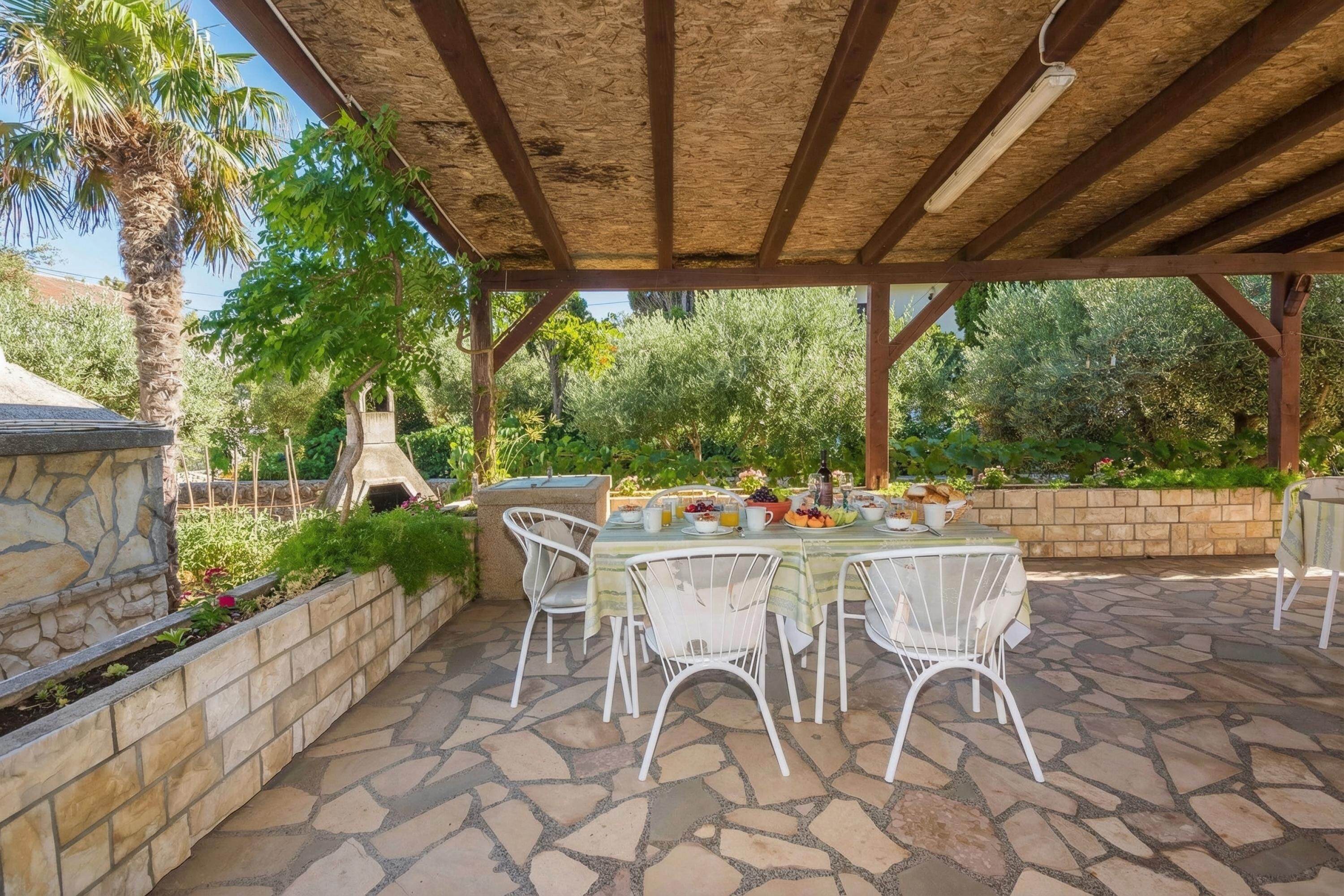 An outdoor patio with stone flooring, white metal chairs, a table set with food, and a pergola overhead.