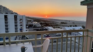 Ein Balkon mit Tisch und Stühlen, der auf einen Parkplatz und einen Strand bei Sonnenuntergang blickt.