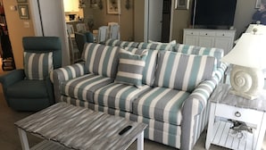 Ein Wohnzimmer mit einem gestreiften Sofa, einem grünen Sessel, einem Holztisch und einem weißen Beistelltisch mit einer Lampe.