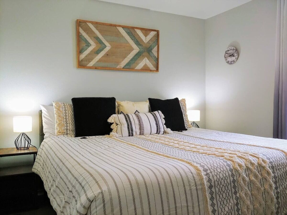 A neatly made bed with striped bedding and decorative pillows, a wall clock, and a geometric artwork above the bed.