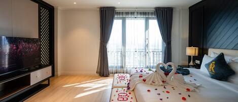 A modern hotel room with a large flat-screen TV, a bed with a heart-shaped towel arrangement, and a balcony with curtains.