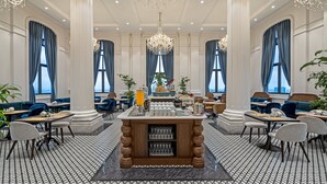 Een ruime hotelentree met een bar in het midden, grote ramen met blauwe gordijnen en een kroonluchter.