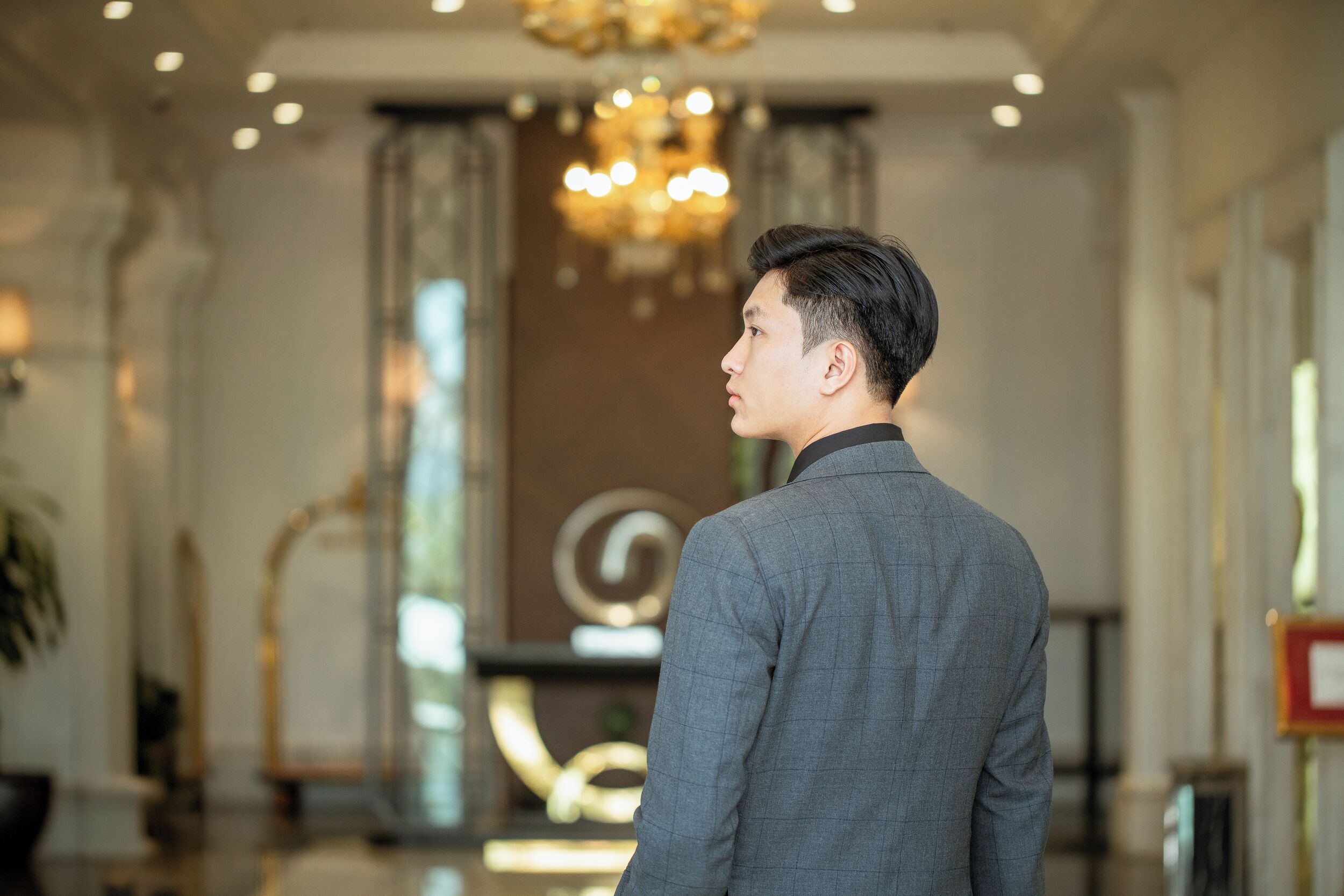 Un hombre con traje gris de pie en un vestíbulo amplio y luminoso, con candelabro, ventanas grandes y una decoración de estilo moderno.