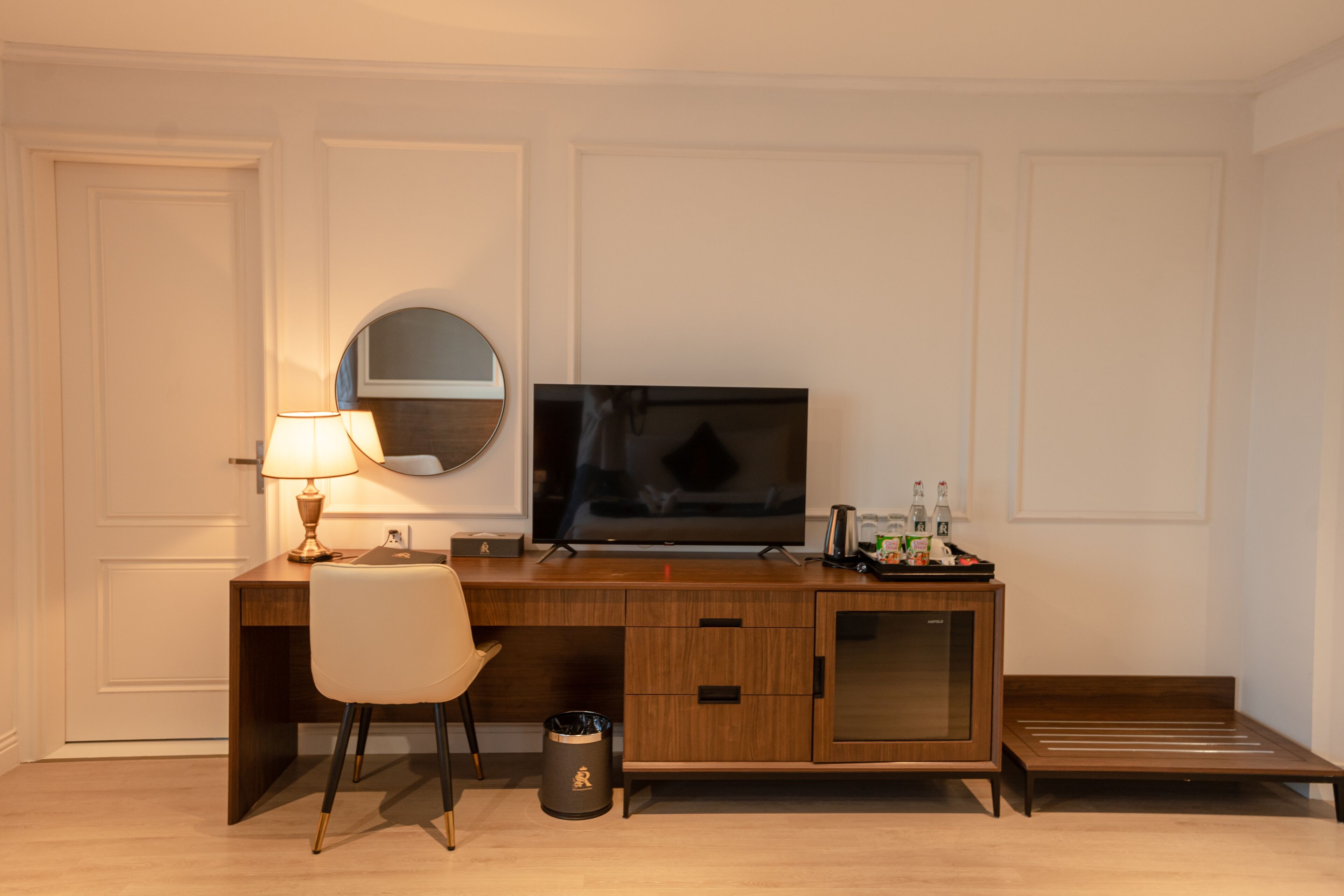Ein Hotelzimmer mit einem Holztisch, einem Fernseher, einer Lampe, einem Stuhl, einem Papierkorb und einem kleinen Schrank.