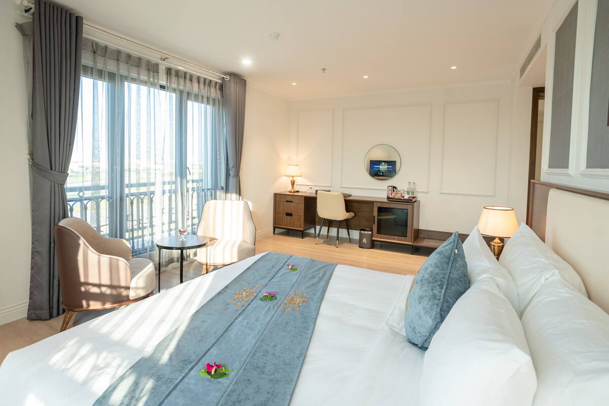 Een hotelkamer met een bed, een bureau, een stoel, een tafeltje en een balkon met gordijnen.