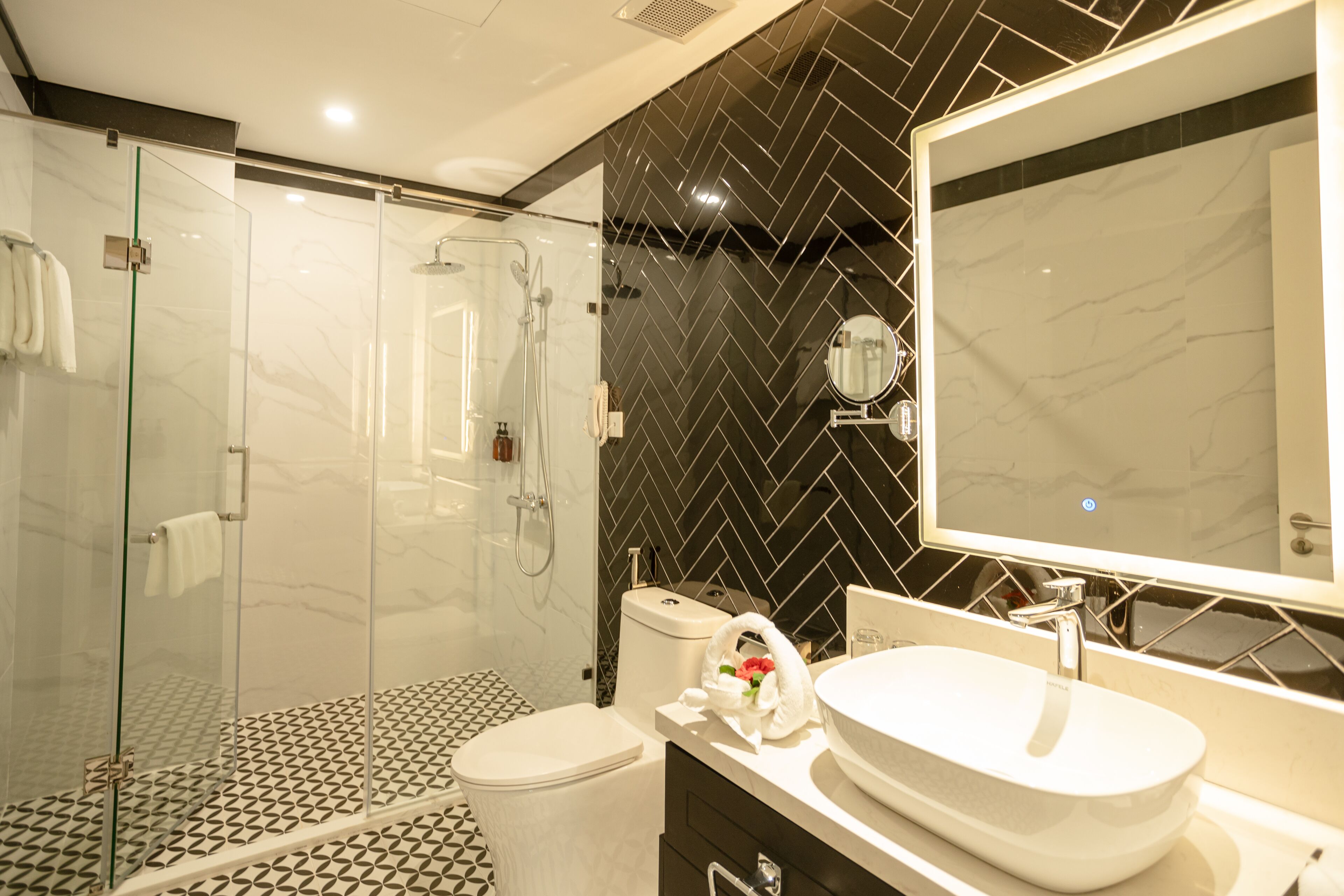 Uma casa de banho moderna com chuveiro em cabine de vidro, lavatório branco e parede com padrão herringbone em preto.