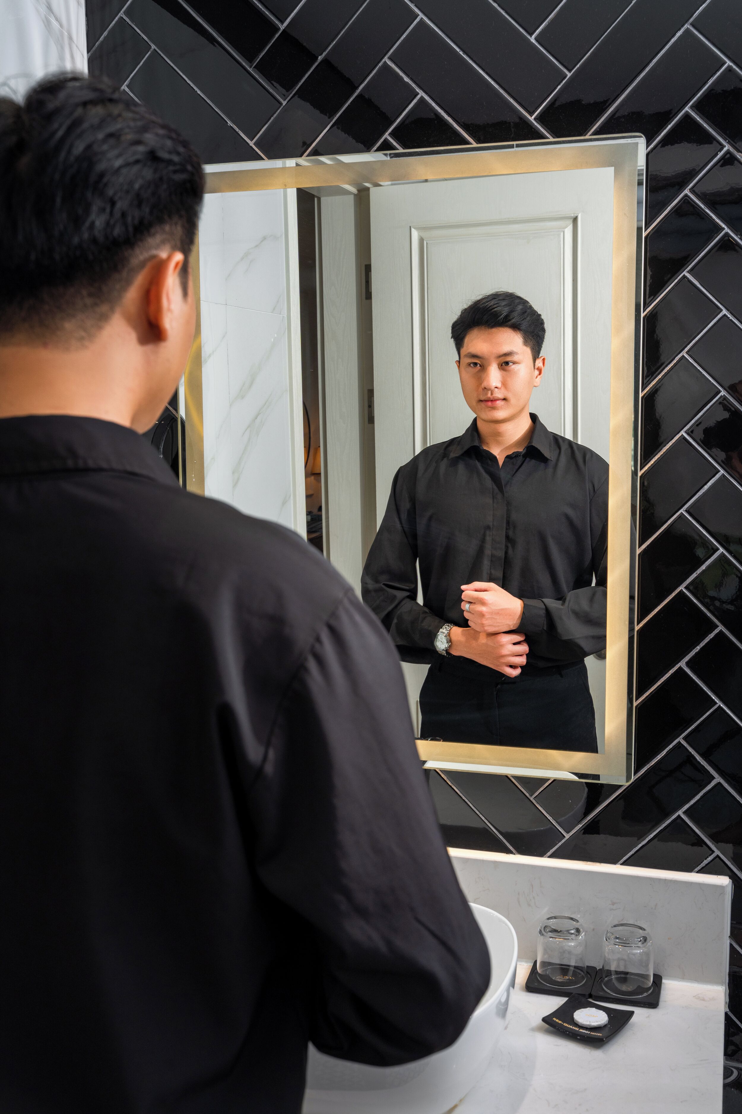 A man in a black shirt standing in front of a bathroom mirror with a white sink and two salt shakers on the counter.