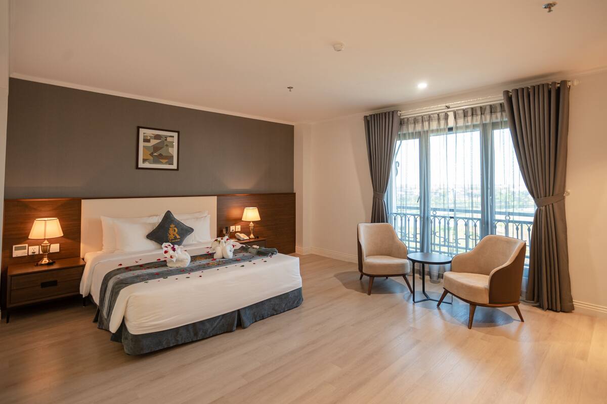 Een hotelkamer met een bed, twee nachtkastjes met lampen, een stoel, een kleintje tafeltje en een raam met gordijnen.