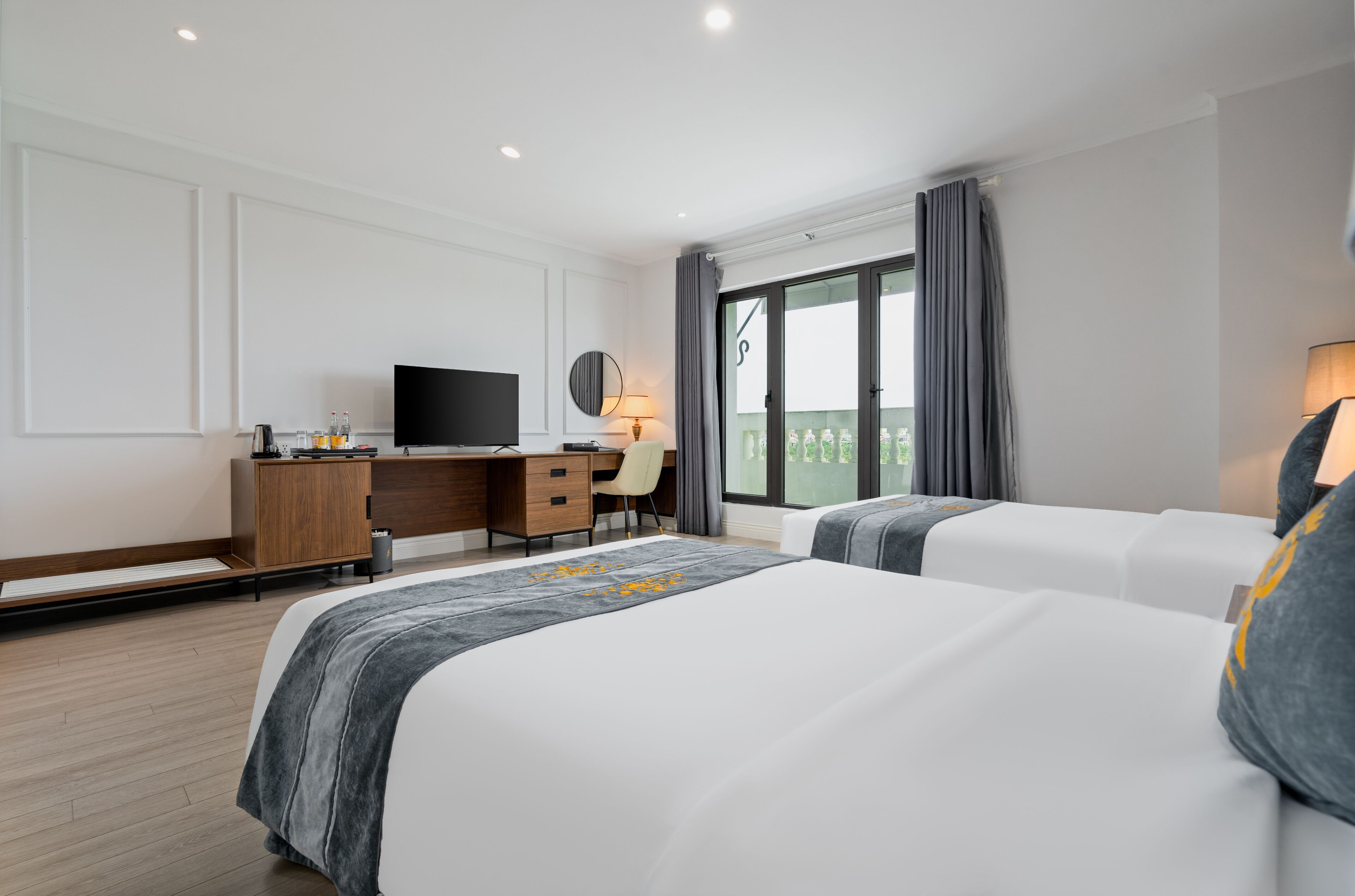 A modern hotel room with two beds, a desk, a TV, and a balcony with curtains.